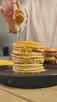 Generous dollop of maple syrup or honey falls from spoon onto plate of freshly prepared, mouthwatering banana-flavored pancakes, topped with berries, on wooden table in dining area. video