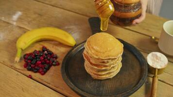 Large drop of maple syrup or honey drips from spoon onto plate of freshly baked, delicious banana-flavored pancakes, with berries, on wooden table in eatery. video