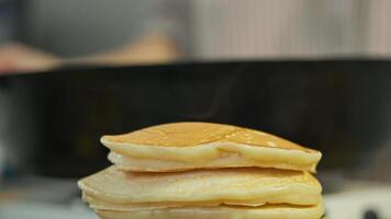 The first stack of hot pancakes is still steaming, while the cook is baking the second batch in the background. Close up. macro video