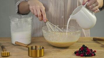 pastry chef generously adds milk to the mixture of flour and eggs while creating a berry pie or pancakes with blueberries. and then whips resulting mixture with whisk by hand in a rustic kitchen. video