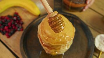 Close-up shows rotating honey dipper, from which maple syrup or honey appetizingly drips onto stack of pancakes surrounded by berries, which creates atmosphere of traditional American breakfast. video
