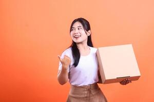 A happy young woman holding a cardboard box, pointing to the left and looking that way. She is smiling, and she seems excited. isolated on orange background photo