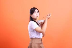 A young woman is captured enthusiastically eating noodles from a bowl with a fork, showcasing a delightful moment of enjoying her meal. isolated on orange background photo