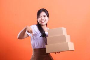A cheerful woman holds a stack of three cardboard boxes and points down while smiling. isolated on orange background photo