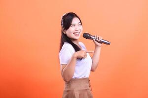 A cheerful young woman holds a microphone, smiling and pointing, ready to perform with passion. isolated on orange background photo