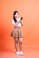 Young smiling Asian woman using smartphone in studio looking happy reading a funny message. isolated on orange background photo