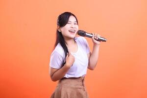A happy asian woman is singing into a microphone with an expressive and joyful look, isolated on orange background photo