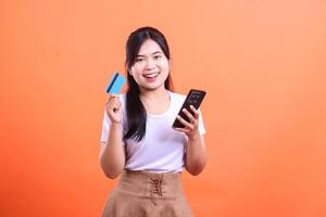 Smiling young woman holding a smartphone and a credit card, looking at the camera with a happy expression. isolated on orange background photo