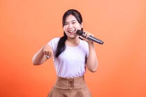 A happy young woman is holding a microphone and pointing down while singing with a joyful expression. isolated on orange background photo