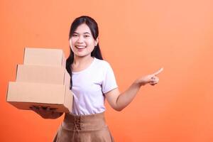 A smiling woman is carrying three stacked cardboard boxes while pointing with her other hand towards the side, isolated on orange background photo