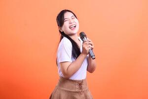 A young woman passionately sings into a microphone, her face radiating happiness and joy with an open mouth, isolated on orange background photo
