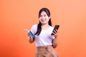 Young cheerful woman showing a credit card and holding a mobile phone, she's smiling and pointing with her finger towards the card, isolated on orange background photo