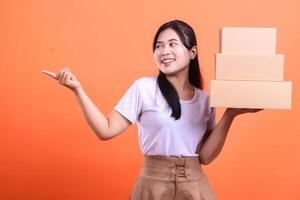 A happy delivery woman holds stack of cardboard boxes while pointing to the side, showing an offer, she is looking toward the direction she is pointing. isolated on orange background photo