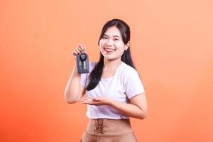 A smiling Asian woman holding out a car key and presenting it with an open palm gesture. isolated on orange background. photo