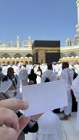 A pilgrim holds a small notebook with a heart doodle in front of the Kaaba in Mecca, Saudi Arabia. The sacred site is surrounded by worshippers performing Hajj or Umrah under a clear blue sky photo