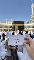 A pilgrim holds a small notebook with a heart doodle in front of the Kaaba in Mecca, Saudi Arabia. The sacred site is surrounded by worshippers performing Hajj or Umrah under a clear blue sky photo