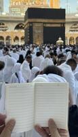 A pilgrim holds a small notebook with a heart doodle in front of the Kaaba in Mecca, Saudi Arabia. The sacred site is surrounded by worshippers performing Hajj or Umrah under a clear blue sky photo