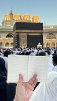 A pilgrim holds a small notebook with a heart doodle in front of the Kaaba in Mecca, Saudi Arabia. The sacred site is surrounded by worshippers performing Hajj or Umrah under a clear blue sky photo