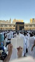 A pilgrim holds a small notebook with a heart doodle in front of the Kaaba in Mecca, Saudi Arabia. The sacred site is surrounded by worshippers performing Hajj or Umrah under a clear blue sky photo