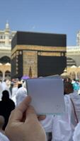 A pilgrim holds a small notebook with a heart doodle in front of the Kaaba in Mecca, Saudi Arabia. The sacred site is surrounded by worshippers performing Hajj or Umrah under a clear blue sky photo