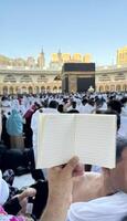 A pilgrim holds a small notebook with a heart doodle in front of the Kaaba in Mecca, Saudi Arabia. The sacred site is surrounded by worshippers performing Hajj or Umrah under a clear blue sky photo