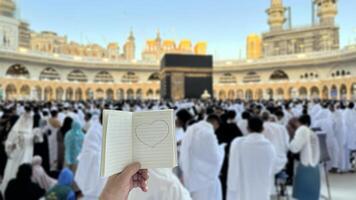 A pilgrim holds a small notebook with a heart doodle in front of the Kaaba in Mecca, Saudi Arabia. The sacred site is surrounded by worshippers performing Hajj or Umrah under a clear blue sky photo