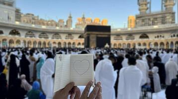A pilgrim holds a small notebook with a heart doodle in front of the Kaaba in Mecca, Saudi Arabia. The sacred site is surrounded by worshippers performing Hajj or Umrah under a clear blue sky photo