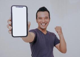 Portrait of excited asian man holding a smartphone with white blank screen in hand, showing close to camera and clenching fist. Gadget with empty free space for mock up and banner, isolated on white. photo