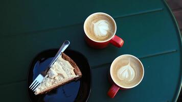 Sweet meringue piece of cake with airy caramel cream and chocolate on a plate in a cafe. 2 aromatic latte coffee in beautiful cups with latte art on a metal table. video