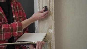 Person measuring a wall with a laser distance meter while taking notes in a notebook. Home renovation, construction planning, and professional measurement accuracy video