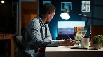 African American freelancer working from home writing notes and researching data driven topics. Focused on productivity and planning tasks in a modern apartment for remote work success. Camera A. video