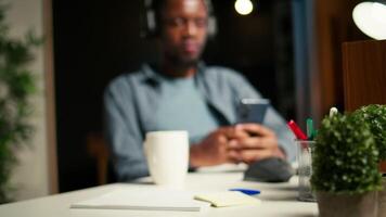 African american person takes a break from work to answer texts at home, using his smartphone to scroll social media apps. Young guy enjoying remote job at home, listens to music on phone. Camera A. video