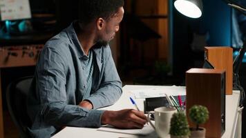 African American freelancer working from home writing notes and researching data driven topics. Focused on productivity and planning tasks in a modern apartment for remote work success. Camera B. video