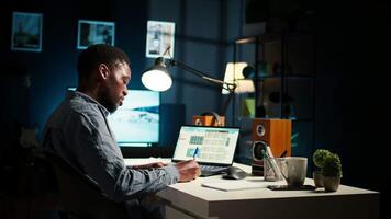 African American teleworker managing daily schedule and does post it memo notes, creating a reminder to ensure task completion from his home office. Freelancer improving productivity. Camera A. video
