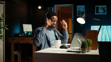 Young casual guy listening to a podcast while he works on freelance job at home, enjoys multitasking with headphones. Man solving tasks and paying attention to webinar discussion. Camera A. video