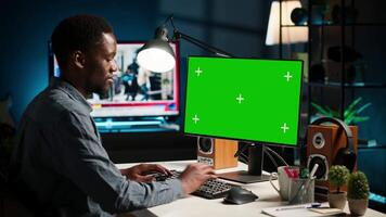 African american person working from home and checking green screen on PC, enjoys remote job at a home office setup. Teleworker multitasking and networking in his modern loft. Camera B. video