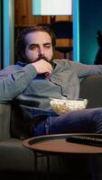 Vertical Homeowner bored at home munching on snacks, using TV to binge series on subscription based streaming services. Man sitting on couch, wasting time with VOD shows on TV, eating popcorn, camera A video