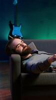 Vertical Smiling man lounged on couch at home eating popcorn while browsing though movies available on streaming service, enjoying time off from work. Man laying down in apartment consuming snacks, camera A video