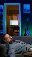 Vertical Exhausted man sleeping on couch after working on laptop all day before managing to watch TV and eat popcorn. Freelancer falls asleep while solving tasks on notebook and watching film, camera B video