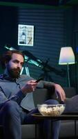 Vertical Man sitting on couch browsing though movies available on streaming service, enjoying time off from work. Person in apartment using remote control to switch channel on television set, camera B video
