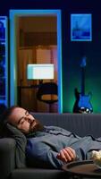Vertical Sleepy man struggling to stay awake on couch after working on laptop all day before managing to watch TV and eat popcorn. Person falls asleep while solving tasks on notebook and watching film video