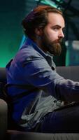 Vertical Man working on laptop lounged on neon lit apartment couch while picking TV channel to watch as distraction. Remote worker solving company tasks on notebook with TV show playing in background, camera A video