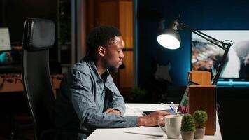 Focused remote worker balancing freelance tasks and researching data online, drafting notes and information for articles. Black person working at home, planning schedule and projects. Camera B. video