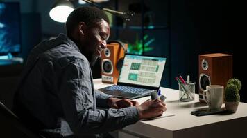 Black freelancer researching online data and writing notes for articles, sitting at his personal home office. Young man working from home, balancing tasks using a wireless setup. Camera B. video