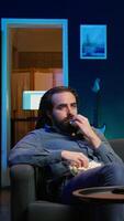 Vertical Sad man on couch eating popcorn from stress while watching heartbreaking drama movie on streaming service. Person consuming snack, distressed by TV show favorite character dies, camera B video