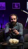 Vertical Excited man playing videogames using controller, having fun in neon illuminated room. Enthusiastic gamer competing in multiplayer game using gamepad, talking with friends through headset, camera B video