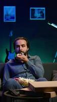 Vertical Exhausted man watching shows on TV late at night begore going to sleep and eating popcorn. Sleepy man in pajamas eating snack on couch, consuming mindless entertainment, camera B video
