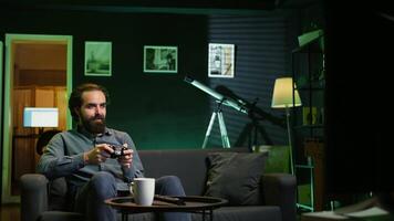 Joyful gamer at home gaming and enjoying leisure time. Smiling man sitting on couch in neon lit living room, excited to play videogame on console, relaxing after hard day at work, camera B video