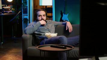 Homeowner bored at home munching on snacks, using TV to binge series on subscription based streaming services. Man sitting on couch, wasting time with VOD shows on TV, eating popcorn, camera A video