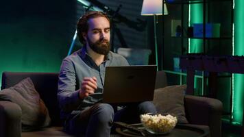Man typing on laptop keyboard in neon lit apartment, sitting on couch and watching TV with bowl of popcorn ready. Teleworking person solving tasks on notebook with film playing in background, camera A video